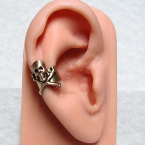 May include: A gold-toned ear cuff featuring a skull and crossbones design. The cuff is shown on a model ear.