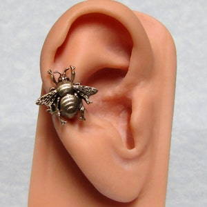 May include: A bronze-colored bee-shaped earring. The earring is highly detailed, with visible wings, body segments, and antennae. The bee is attached to a person's ear, showcasing the earring's size and design. The earring is a unique piece of jewelry.