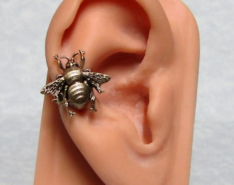 May include: A bronze-colored bee-shaped earring. The earring is highly detailed, with visible wings, body segments, and antennae. The bee is attached to a person's ear, showcasing the earring's size and design. The earring is a unique piece of jewelry.