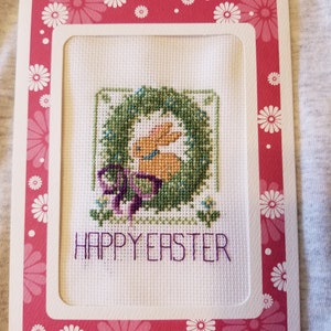 May include: A white card with a pink border featuring a white daisy pattern. The card has a white inner frame with a cross-stitch design of a bunny rabbit inside a green wreath. The text "HAPPY EASTER" is stitched in purple below the bunny.