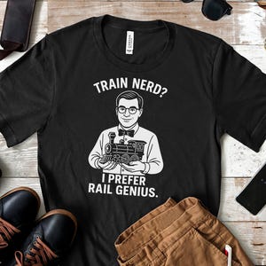 May include: Black t-shirt with a white graphic of a man holding a train, with the text "TRAIN NERD? I PREFER RAIL GENIUS." The shirt is surrounded by accessories, including shoes and a phone.