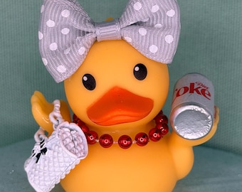 May include: A yellow rubber duck featuring a grey polka-dot bow on its head, a red beaded necklace, a white quilted handbag on one wing, and a miniature silver Diet Coke can held in the other wing. The duck has black eyes and an orange beak.