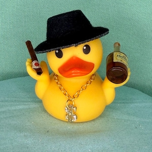 Könnte beinhalten: Eine gelbe Gummiente mit einem schwarzen Fedora-Hut und einer goldfarbenen Kette mit einem Dollarzeichen-Anhänger. Die Ente hält eine braune Zigarre in ihrem rechten Flügel und eine kleine braune Flasche mit einem Etikett im linken Flügel.