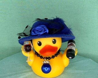 May include: A yellow rubber duck wearing a blue felt hat with a black veil and a blue fabric rose. The duck wears a black beaded necklace with a large blue pendant and holds a miniature glass of red wine and a miniature bottle of red wine.