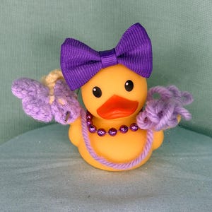May include: A yellow rubber duck featuring a purple ribbon bow on its head, a purple beaded necklace, and light purple crocheted yarn accents attached to its sides. The duck has a bright orange beak and black eyes, creating a colorful appearance.