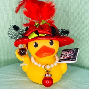 May include: A yellow rubber duck wearing a red hat with red feathers, green ribbons, and black netting. It wears a white pearl necklace with a red ornament pendant. The duck holds a miniature wine glass and a book titled A Good Girl's Guide to Murder.
