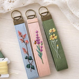 Könnte beinhalten: Drei Schlüsselanhänger aus Stoff mit gestickten Blumenmotiven. Der hellblaue Anhänger zeigt einen rosa Blütenzweig, der rosafarbene zeigt lila Lavendel mit einer kleinen Biene und der olivgrüne zeigt gelbe Blumen. Jeder hat einen Schlüsselring aus bronzefarbenem Metall.
