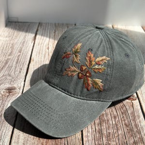 Op de afbeelding: Een grijze baseballcap met een gebogen klep. De cap heeft een geborduurd ontwerp van herfstbladeren en eikels in bruin, groen en oranje tinten. De hoed is gemaakt van een zachte, getextureerde stof.
