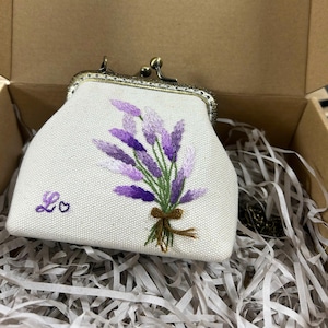 Op de afbeelding: Een witte, geborduurde portemonnee met een metalen sluiting. De portemonnee heeft een lavendelboeket met paars en groen garen, vastgebonden met een bruine strik. De initialen "Lo" en een hartje zijn op de voorkant geborduurd. De portemonnee zit in een kartonnen doos.