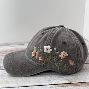 Op de afbeelding: Een grijze baseballcap met geborduurde bloemen in roze, wit en bruin. De cap heeft een gebogen klep en een knoop aan de bovenkant. De bloemenborduring zit aan de linkerkant.