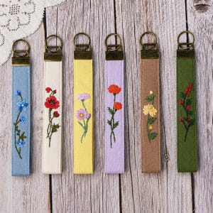 Könnte beinhalten: Sechs Schlüsselanhänger aus Stoff mit gestickten Blumenmotiven in Blau, Weiß, Gelb, Lavendel, Braun und Grün. Jedes Band verfügt über einen bronzefarbenen Metallverschluss und einen Ring mit detaillierten botanischen Stickereien auf einfarbigem Stoff.