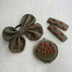 May include: An olive green hair bow, two barrettes, and a compact mirror. Each item is decorated with small, embroidered red flowers. The bow is attached to a black elastic band. The compact mirror has a floral design.