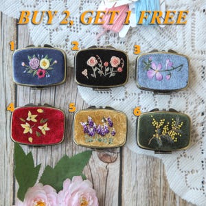 Op de afbeelding: Zes rechthoekige fluwelen dozen met geborduurde bloemmotieven. De dozen zijn in verschillende kleuren, waaronder marineblauw, zwart, lichtblauw, rood, goud en groen. De tekst "BUY 2, GET 1 FREE" staat bovenaan de afbeelding.