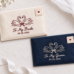Puede incluir: Dos sobres rectangulares de tela con diseños de cisnes bordados. El sobre crema dice To My Bride 09.25.2026 con un sello de corazón rojo. El sobre azul marino dice To My Groom 09.25.2026 con un sello de corazón rojo y lazo crema.