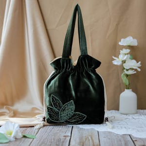 Puede incluir: Un bolso con cordón de terciopelo verde oscuro con asas a juego. El bolso presenta un diseño de hojas bordadas con detalles de cuentas. El bolso está sobre una superficie de madera con flores blancas y un jarrón en el fondo.
