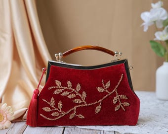 May include: A red velvet handbag with a gold beaded floral design. The bag has a tortoiseshell handle and a red tassel. The frame is black with silver accents. The bag is approximately 10 inches wide and 7 inches tall.