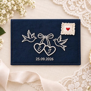 Puede incluir: Una cubierta rectangular de tela azul marino con bordados blancos de dos pájaros volando, un lazo y dos marcos en forma de corazón con las letras E y J. Se observa un pequeño parche cuadrado de encaje crema con un corazón rojo y la fecha 25.09.2026.