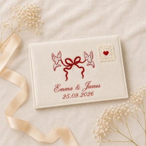 Puede incluir: Un libro de visitas rectangular de tela color hueso con palomas rojas bordadas unidas por un lazo, un pequeño parche de encaje cuadrado con un corazón rojo y el texto bordado Emma & James con la fecha 25.09.2026 en hilo rojo.