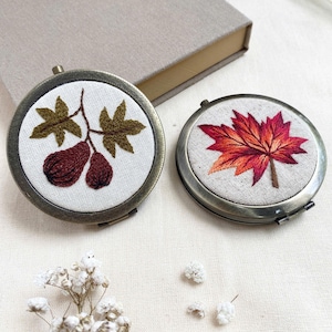 Peut inclure: Deux miroirs compacts ronds de style antique avec des motifs brodés. L'un présente une branche avec des fruits bruns et des feuilles vertes, l'autre une feuille d'érable rouge et orange. Ils reposent sur une surface crème.