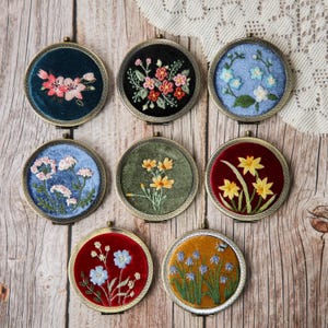 Può includere: Una collezione di sette specchietti compatti rotondi ricamati. Ogni specchietto presenta un diverso disegno floreale su sfondo di velluto, con fiori rosa, rossi, gialli e blu. Gli specchietti hanno una cornice in metallo color bronzo e sono disposti su una superficie di legno.