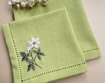 May include: Two square, light green linen napkins. One napkin features an embroidered white flower with green leaves. The edges of the napkins have a decorative, dotted border. A sprig of white flowers is in the background.