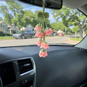 Könnte beinhalten: Ein handgefertigter Autoanhänger mit gehäkelten rosa Blüten mit grünen Stielen und einer grünen Schleife. Der Anhänger hängt am Rückspiegel eines Autos und verleiht dem Innenraum eine handgefertigte Dekoration.