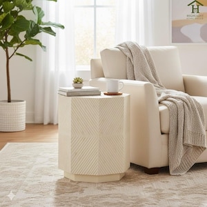 May include: A cream-coloured, geometric side table with a hexagonal shape. The table has a textured surface and is paired with a cream-coloured armchair, a knitted throw, and a patterned rug. A small plant and a mug sit on top of the table.