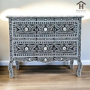 May include: A black and white floral patterned chest of drawers. The chest has two drawers with white flower-shaped knobs. The design features a floral and leaf pattern. The chest has curved legs and a decorative edge.