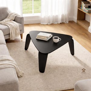 Könnte beinhalten: Ein schwarzer, dreieckiger Couchtisch mit drei gebogenen Beinen. Der Tisch hat eine matte Oberfläche und trägt einen Stapel Bücher und eine weiße Kaffeetasse. Der Tisch steht auf einem hellen Teppich in einem Wohnzimmer.