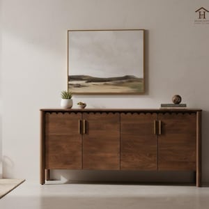 Può includere: Una credenza in legno marrone scuro con quattro ante e maniglie in ottone. Il bordo superiore ha un design smerlato. Sopra la credenza è appeso un dipinto di paesaggio incorniciato e sopra ci sono una piccola pianta e oggetti decorativi.