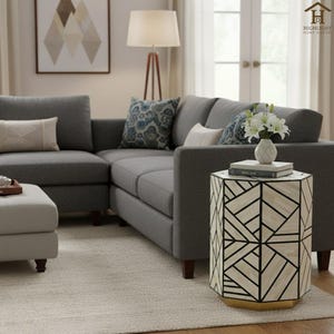 Könnte beinhalten: Ein modernes Wohnzimmer mit einem grauen Ecksofa, einem gemusterten Beistelltisch und einem beigen Hocker. Der Beistelltisch hat ein geometrisches Design mit schwarzen Linien und einem goldenen Sockel. Eine Lampe und Kunstwerke sind ebenfalls zu sehen.