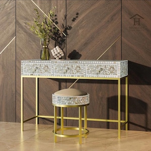 Könnte beinhalten: Ein dekoratives Schminktisch- und Hocker-Set. Der Tisch hat drei Schubladen mit floralen Designs und goldfarbenen Beinen. Der passende Hocker hat einen gepolsterten Sitz und ein ähnliches Blumendesign. Eine Vase mit Grün steht auf dem Tisch.