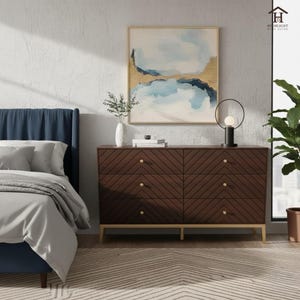 Könnte beinhalten: Schlafzimmer-Interieur mit einer dunkelbraunen Kommode mit sechs Schubladen und goldfarbener Hardware. Die Schubladen haben ein Fischgrätmuster. Ein blaues Bett und abstrakte Kunst mit goldenen Akzenten sind ebenfalls zu sehen. Eine Lampe und eine Pflanze vervollständigen die Einrichtung.