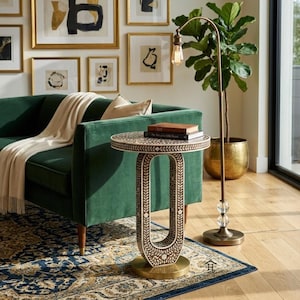 Könnte beinhalten: Ein dekorativer Beistelltisch mit gemusterter Tischplatte und ovalem Sockel neben einem grünen Samtsofa. Der Tisch hat einen goldfarbenen Sockel und ist mit einem floralen Design verziert. Eine Stehlampe mit Vintage-Glühbirne und eine Topfpflanze befinden sich ebenfalls im Raum.