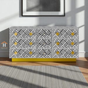 Könnte beinhalten: Eine moderne Kommode mit sechs Schubladen und einem schwarz-weißen geometrischen Muster. Die Kommode hat goldfarbene Knöpfe und einen leuchtend gelben Sockel. Das Möbelstück steht vor einer neutralen Wand mit gerahmter Kunst und einem grauen Teppich.