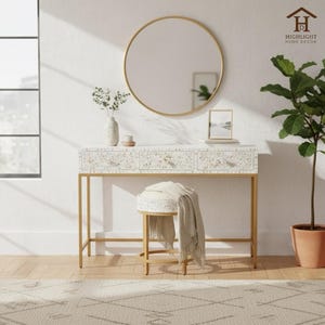 Könnte beinhalten: Ein Schminktisch mit goldenem Rahmen und einer weißen, floral gemusterten Oberseite mit drei Schubladen. Ein runder goldener Spiegel befindet sich darüber. Ein passender Hocker mit Decke und eine Topfpflanze vervollständigen die Szene. Der Text "HIGHLIGHT HOME DECOR" ist sichtbar.