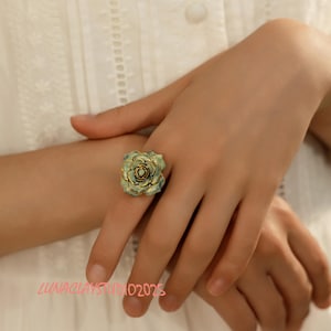 May include: A handcrafted ring featuring a detailed, three-dimensional rose design. The ring has a teal and gold color scheme, with the rose petals in teal and the edges highlighted in gold. The ring is worn on a finger, with the text "LUNACLAYS STUDIO 02023" visible.