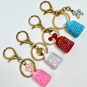 May include: Four keychains with gold-tone hardware and decorative charms. Each keychain features a small, bedazzled baby bottle charm in pink, white, red, and blue. Additional charms include a flower, cherries, a heart, and a floral design.