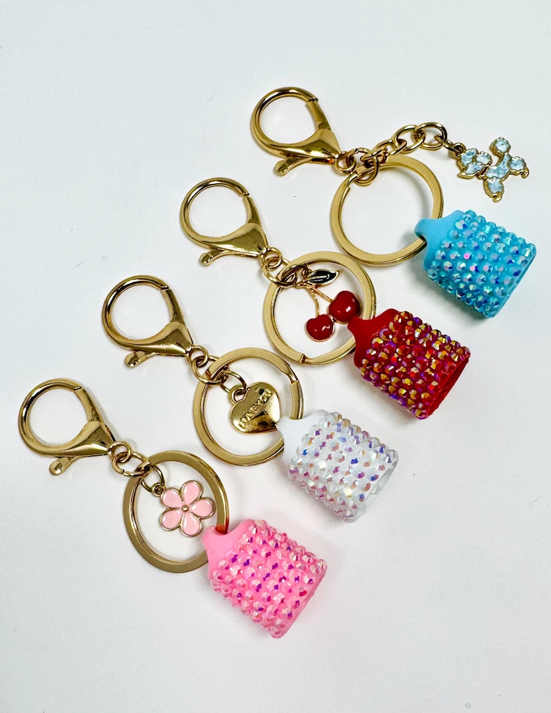May include: Four keychains with gold-tone hardware and decorative charms. Each keychain features a small, bedazzled baby bottle charm in pink, white, red, and blue. Additional charms include a flower, cherries, a heart, and a floral design.