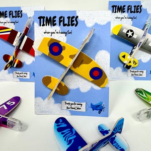 May include: Airplane-shaped party favors with the text "TIME FLIES when you're having fun!" on blue cards. The favors are in various colors and designs, including yellow, red, and blue. Each airplane has a pen attached.