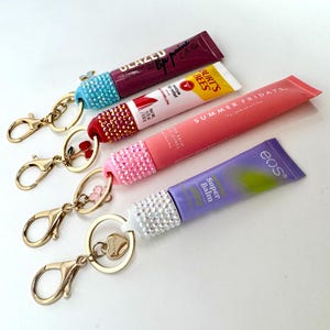 May include: Four lip balm tubes with rhinestone-studded caps attached to gold-colored keychains. The tubes are various colors, including purple, pink, and yellow. One keychain has a heart charm, and another has a flower charm.