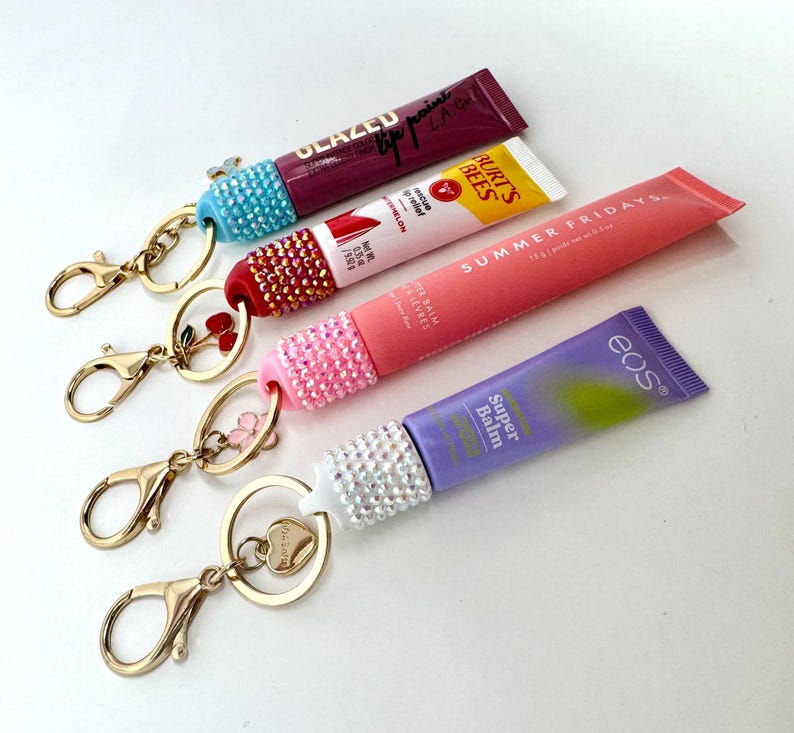 May include: Four lip balm tubes with rhinestone-studded caps attached to gold-colored keychains. The tubes are various colors, including purple, pink, and yellow. One keychain has a heart charm, and another has a flower charm.
