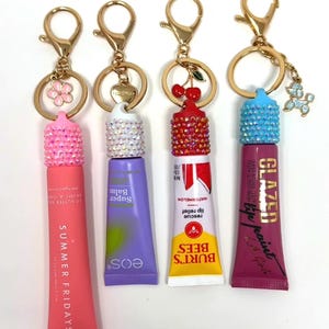 May include: Four lip balm keychains with gold-tone hardware. The lip balms are in pink, purple, white and red tubes. The keychains have decorative charms, including a flower, a heart, cherries, and a poodle.