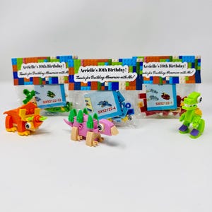 May include: A collection of colorful toy dinosaur figures and building block sets, with the text "Arrielle's 10th Birthday! Thanks for Building Memories with Me!" on the packaging. The toys are in orange, pink, green, and blue.