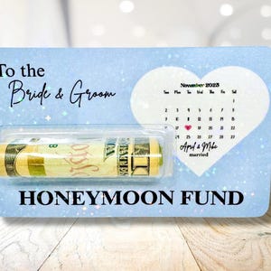 May include: A light blue gift card with the text "To the Bride & Groom" and "Honeymoon Fund". It features a rolled-up bill and a calendar with a heart on the wedding date.