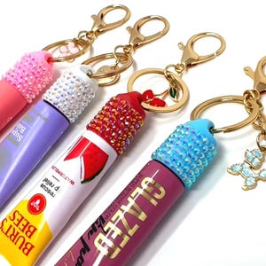 May include: A collection of lip gloss keychains in various colors, including pink, purple, red, and magenta. Each tube is adorned with sparkling rhinestones on the cap and attached to a gold-colored keychain. Some keychains feature charms.