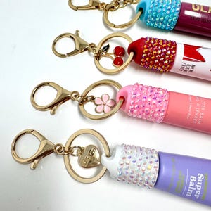 May include: Four lip balm keychains with gold-colored hardware. Each keychain features a different colored lip balm tube, adorned with rhinestones. The charms include a cherry, a flower, a heart, and a small cross.