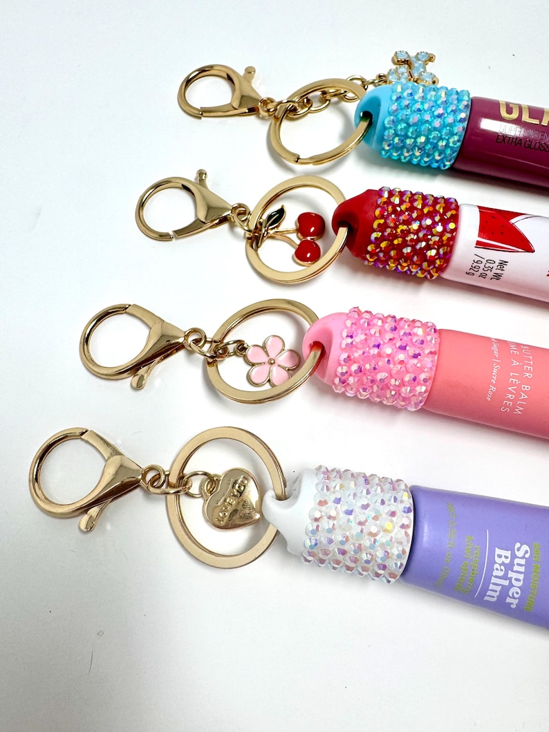 May include: Four lip balm keychains with gold-colored hardware. Each keychain features a different colored lip balm tube, adorned with rhinestones. The charms include a cherry, a flower, a heart, and a small cross.
