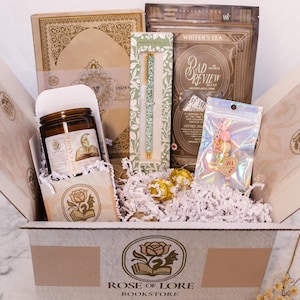Puede incluir: Una caja de regalo de Rose of Lore Bookstore llena de una vela de soja, un diario, un bolígrafo, té y un llavero. La caja contiene papel decorativo y caramelos envueltos en oro. La vela tiene una etiqueta con una ilustración. El té está etiquetado "Bad Review".