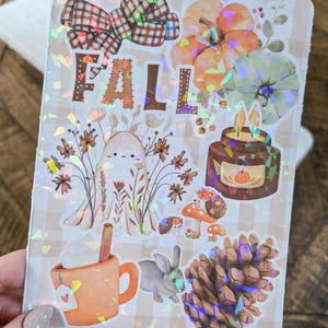 May include: A decorative sticker with a fall theme. The sticker features the word "FALL", pumpkins, a bow, a candle, a bunny, and a pinecone. The background is a light brown and white checkered pattern.